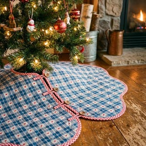 May include: A quilted Christmas tree skirt featuring a blue and white plaid pattern with small red hearts. The circular fabric mat has a scalloped edge trimmed in red and white gingham, secured with four wooden buttons and fabric loops.