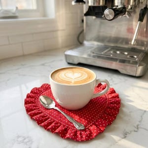 Puede incluir: Una taza de cer&aacute;mica blanca llena de arte latte en forma de coraz&oacute;n, sobre un posavasos rojo en forma de coraz&oacute;n con lunares blancos y un borde con volantes. Una cuchara plateada est&aacute; al lado de la taza.