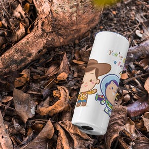 May include: White travel mug with a cartoon image of Woody and Buzz Lightyear from Toy Story. The mug has the text "You're a Friend to Me" printed on it.