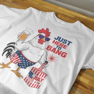 May include: White t-shirt with a cartoon rooster wearing sunglasses and holding a beer. The rooster is wearing red, white, and blue swim trunks and holding a firework. The text "Just Here To Bang" is printed on the shirt.