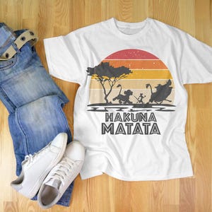 May include: White t-shirt with a colorful sunset design and the phrase "Hakuna Matata". The shirt features a silhouette of characters from The Lion King. The image also includes blue jeans, a belt, and white sneakers.