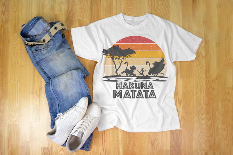 May include: White t-shirt with a colorful sunset design and the phrase "Hakuna Matata". The shirt features a silhouette of characters from The Lion King. The image also includes blue jeans, a belt, and white sneakers.