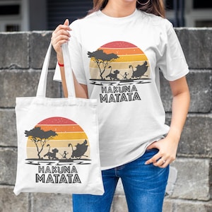 May include: White t-shirt and tote bag featuring a sunset design with silhouettes of characters and the text "HAKUNA MATATA". The design includes a tree and characters from a popular animated film. The tote bag is held by a person.