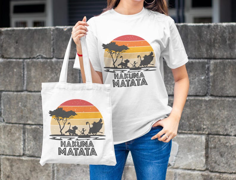 May include: White t-shirt and tote bag featuring a sunset design with silhouettes of characters and the text "HAKUNA MATATA". The design includes a tree and characters from a popular animated film. The tote bag is held by a person.