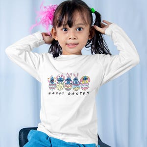 May include: A white long-sleeved shirt with a graphic of blue cartoon characters in colorful Easter eggs. The text "Happy Easter" is printed below the graphic.