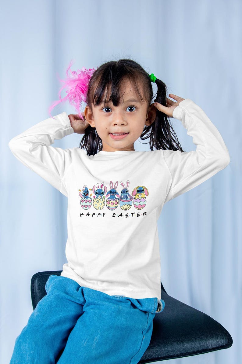May include: A white long-sleeved shirt with a graphic of blue cartoon characters in colorful Easter eggs. The text "Happy Easter" is printed below the graphic.