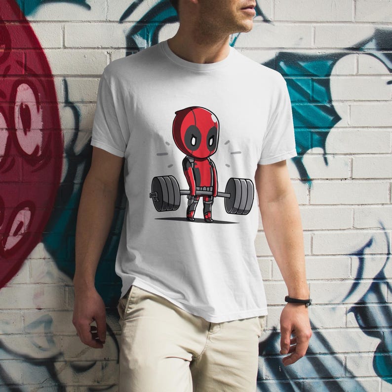 May include: White t-shirt featuring a cartoon image of Deadpool lifting a barbell. The image is in a comic book style with red, black, and white colours.