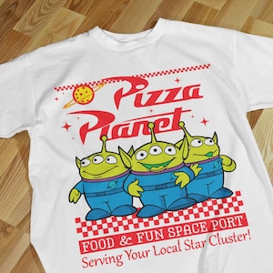 May include: White t-shirt with a graphic of three green aliens from the movie Toy Story. The aliens are standing in front of a red and white checkered background with the text "Pizza Planet" in red and white. The text "Food & Fun Space Port" and "Serving Your Local Star Cluster!" is also printed on the shirt.