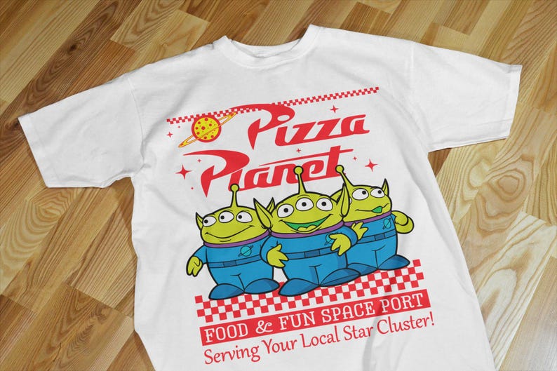 May include: White t-shirt with a graphic of three green aliens from the movie Toy Story. The aliens are standing in front of a red and white checkered background with the text "Pizza Planet" in red and white. The text "Food & Fun Space Port" and "Serving Your Local Star Cluster!" is also printed on the shirt.