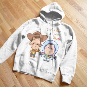 May include: White hoodie with a cartoon image of Woody and Buzz Lightyear from Toy Story. The text "You've got a friend in me" is printed in rainbow colors on the hoodie.