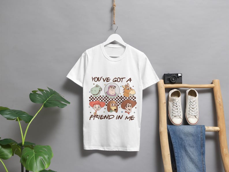 Op de afbeelding: Wit T-shirt met een grafische print van personages uit de Toy Story-filmfranchise. De grafiek toont Woody, Buzz Lightyear, Jessie, Rex en Slinky Dog. De tekst "You've got a friend in me" is onder de grafiek gedrukt.