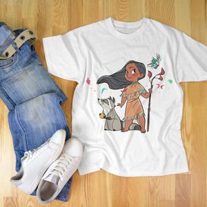May include: White t-shirt with a cartoon illustration of a Native American girl with long black hair, wearing a brown dress and holding a stick. A raccoon is standing next to her. The girl is surrounded by flowers and leaves.