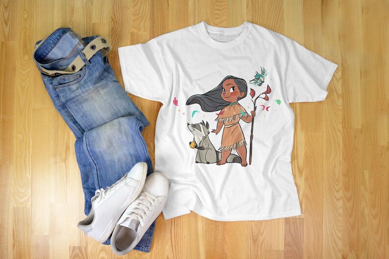 May include: White t-shirt with a cartoon illustration of a Native American girl with long black hair, wearing a brown dress and holding a stick. A raccoon is standing next to her. The girl is surrounded by flowers and leaves.