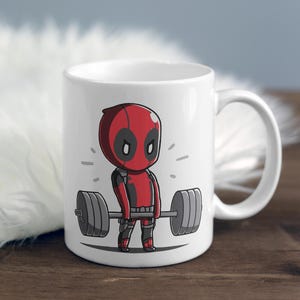May include: White ceramic coffee mug featuring a red and black cartoon character lifting a barbell. The character has a red and black mask and is wearing a black suit. The mug has a black handle.