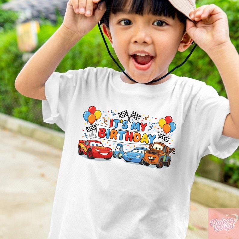 May include: White t-shirt featuring a colorful graphic of animated cars and the text "IT'S MY BIRTHDAY". The design includes balloons and checkered flags, perfect for a birthday celebration.