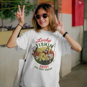 May include: White t-shirt with a retro-style graphic of Goofy fishing. The graphic features a yellow, green, and brown color scheme and the text "Lucky Fishin' Shirt Do Not Wash".