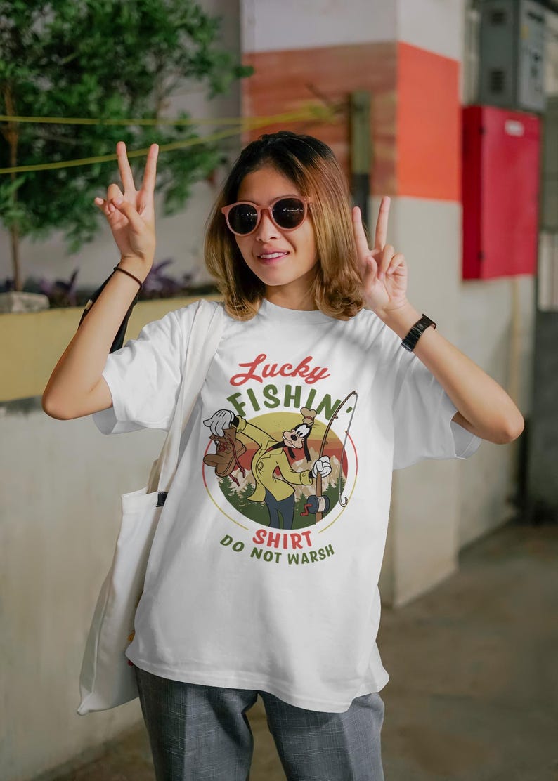 May include: White t-shirt with a retro-style graphic of Goofy fishing. The graphic features a yellow, green, and brown color scheme and the text "Lucky Fishin' Shirt Do Not Wash".