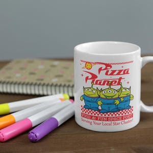 May include: White ceramic mug with a red and white design featuring the words "Pizza Planet" and three green alien characters from the Toy Story movie. The text below the characters reads "Food & Fun Space Port Serving Your Local Star Cluster!"