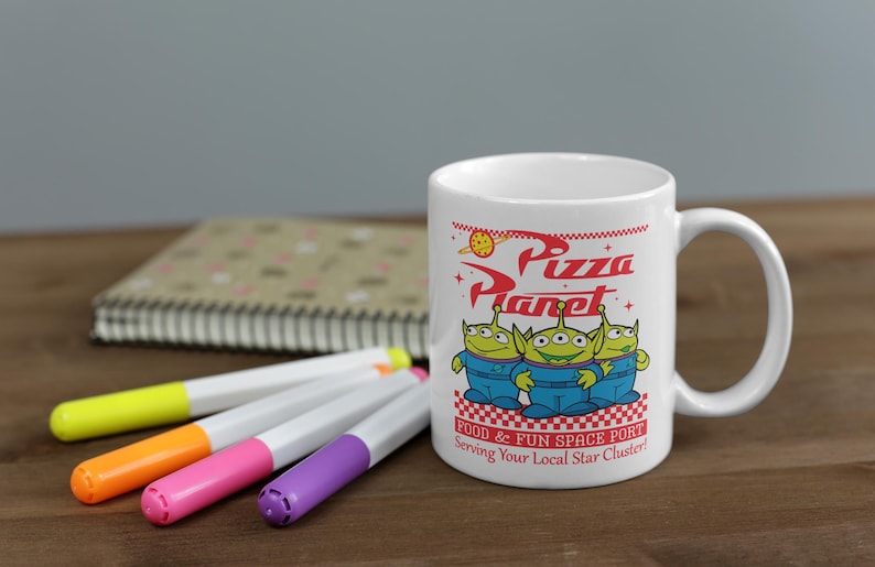 May include: White ceramic mug with a red and white design featuring the words "Pizza Planet" and three green alien characters from the Toy Story movie. The text below the characters reads "Food & Fun Space Port Serving Your Local Star Cluster!"