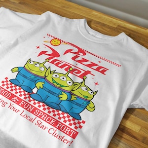 May include: White t-shirt with a graphic of three green aliens from the Toy Story franchise. The aliens are holding hands and standing in front of a red and white checkered background. The text "Pizza Planet" is written in red and white above the aliens. The text "Food & Fun Space Port Serving Your Local Star Cluster!" is written in red and white below the aliens.