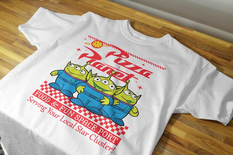 May include: White t-shirt with a graphic of three green aliens from the Toy Story franchise. The aliens are holding hands and standing in front of a red and white checkered background. The text "Pizza Planet" is written in red and white above the aliens. The text "Food & Fun Space Port Serving Your Local Star Cluster!" is written in red and white below the aliens.