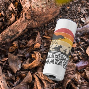 May include: White tumbler with a colorful sunset design and the words "Hakuna Matata." The design includes silhouettes of animals and a tree. The tumbler is cylindrical and sits among fallen leaves and branches.