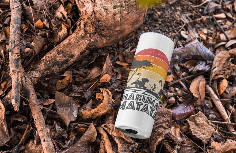 May include: White tumbler with a colorful sunset design and the words "Hakuna Matata." The design includes silhouettes of animals and a tree. The tumbler is cylindrical and sits among fallen leaves and branches.