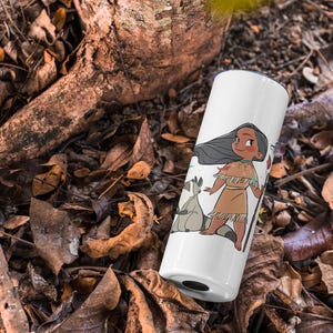 May include: White tumbler with a cartoon illustration of a Native American girl with long black hair, wearing a brown dress and holding a feather. Two white raccoons are beside her. The tumbler is on a bed of brown leaves.