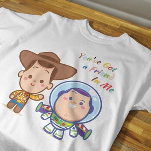 May include: White t-shirt with a cartoon image of Woody and Buzz Lightyear from Toy Story. The shirt has the text "You've Got a Friend in Me" printed on it in rainbow colors.