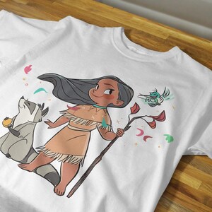 May include: White t-shirt with a cartoon illustration of a Native American girl with long black hair, wearing a brown dress and holding a stick. She is surrounded by a raccoon, a bird, and colorful flowers.