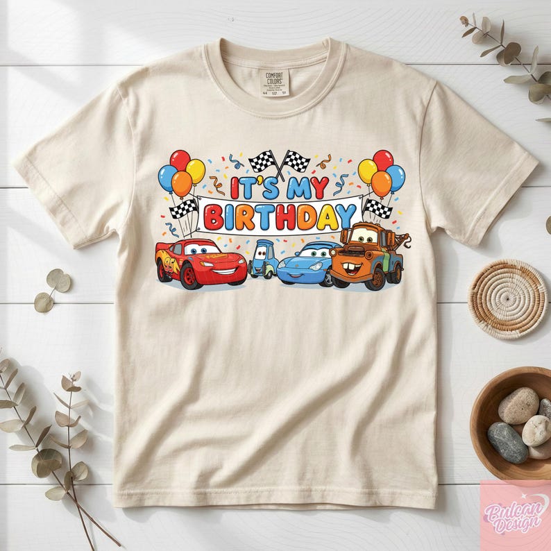May include: A beige t-shirt featuring a colorful graphic of animated cars and the text "IT'S MY BIRTHDAY." The design includes balloons, checkered flags, and confetti. The shirt is made of a soft material.
