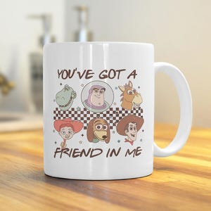 Op de afbeelding: Witte keramische mok met een kleurrijke illustratie van personages uit de Toy Story-franchise. De mok heeft een handvat en de tekst "You've got a friend in me" staat op de voorkant.