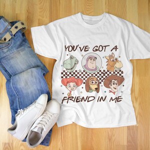 Op de afbeelding: Wit T-shirt met een cartoon afbeelding van personages uit de Toy Story film. Het shirt heeft de tekst "You've got a friend in me" erop gedrukt.