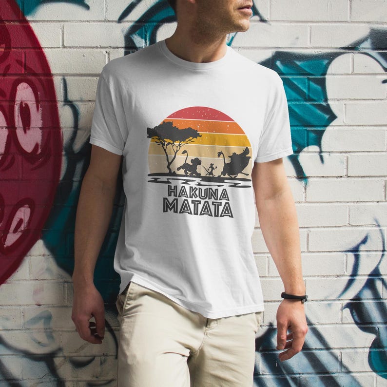 May include: White t-shirt featuring a sunset design with a tree and silhouettes of characters. The phrase "HAKUNA MATATA" is printed in black below the image. The shirt is being worn by a person.