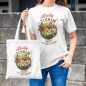 May include: White tote bag with a cartoon character fishing. The bag has the text "Lucky Fishing Shirt Do Not Wash" printed on it.