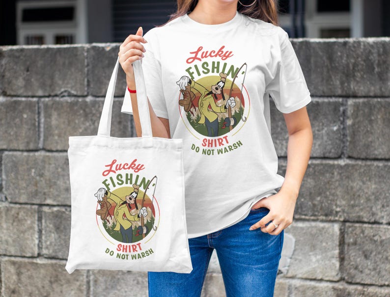 May include: White tote bag with a cartoon character fishing. The bag has the text "Lucky Fishing Shirt Do Not Wash" printed on it.