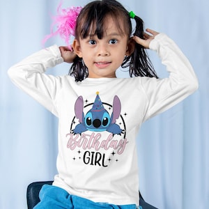 Op de afbeelding: Witte longsleeve met een blauwe cartoonfiguur die een feestmuts draagt. Het figuur kijkt uit een cirkel met sterren en de tekst "Birthday Girl".
