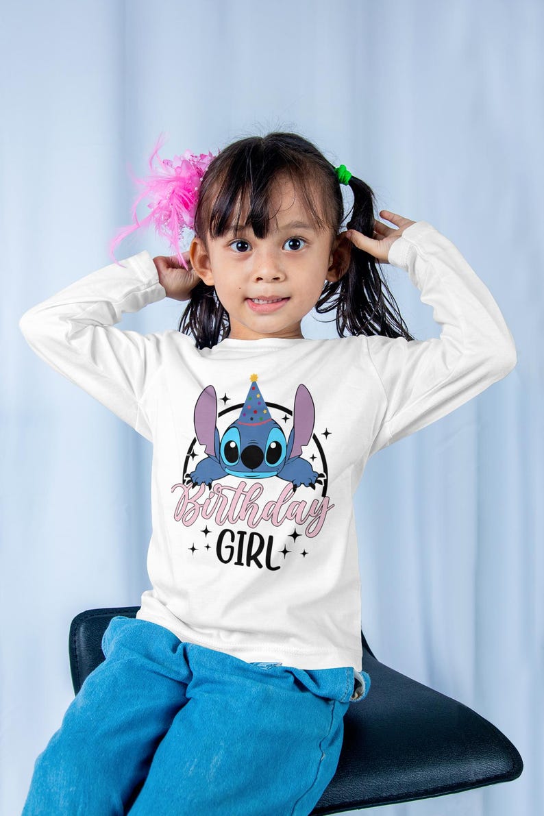 Op de afbeelding: Witte longsleeve met een blauwe cartoonfiguur die een feestmuts draagt. Het figuur kijkt uit een cirkel met sterren en de tekst "Birthday Girl".
