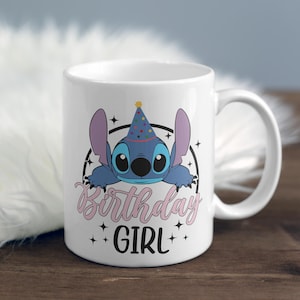 Op de afbeelding: Witte keramische mok met een blauw cartoonfiguur dat een feestmuts draagt. Het figuur kijkt over de rand van de mok. De tekst "Birthday Girl" is in roze onder het figuur gedrukt.