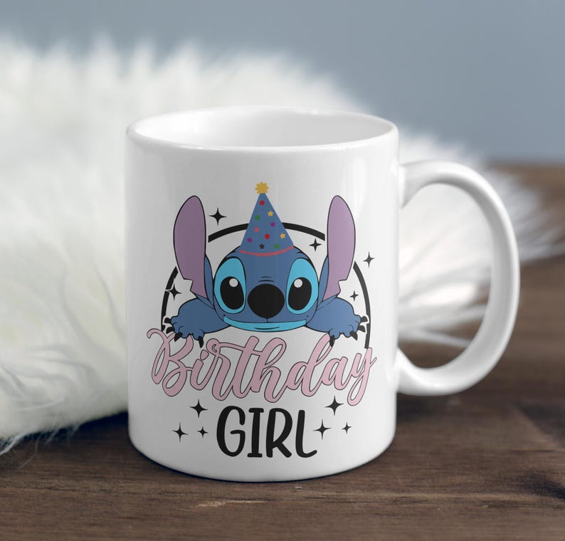 Op de afbeelding: Witte keramische mok met een blauw cartoonfiguur dat een feestmuts draagt. Het figuur kijkt over de rand van de mok. De tekst "Birthday Girl" is in roze onder het figuur gedrukt.