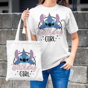 Op de afbeelding: Een witte tote bag met een blauw cartoonfiguur dat een feestmuts draagt en de tekst "Birthday Girl". De tas heeft een zwarte band en wordt vastgehouden door iemand die een wit t-shirt draagt met hetzelfde ontwerp.