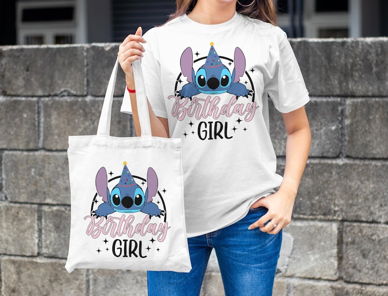 Op de afbeelding: Een witte tote bag met een blauw cartoonfiguur dat een feestmuts draagt en de tekst "Birthday Girl". De tas heeft een zwarte band en wordt vastgehouden door iemand die een wit t-shirt draagt met hetzelfde ontwerp.