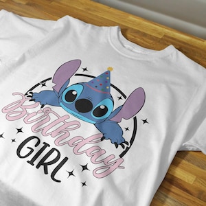 Op de afbeelding: Wit T-shirt met een blauw Stitch-personage dat een feestmuts draagt. De tekst "Birthday Girl" is geschreven in roze en zwart.
