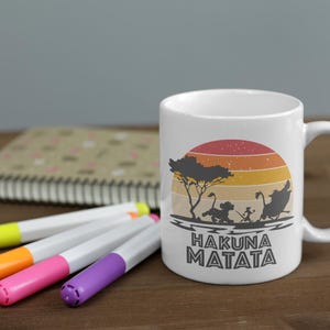 May include: White ceramic mug featuring a sunset design with silhouettes of characters and the text "HAKUNA MATATA". The mug is accompanied by colorful markers and a notebook on a wooden surface.