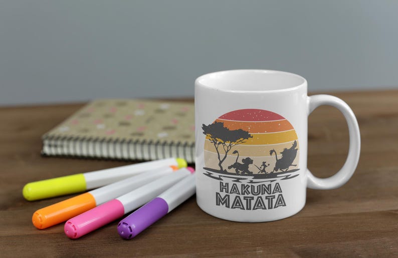 May include: White ceramic mug featuring a sunset design with silhouettes of characters and the text "HAKUNA MATATA". The mug is accompanied by colorful markers and a notebook on a wooden surface.