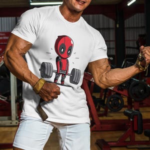 May include: White t-shirt featuring a cartoon image of Deadpool lifting weights. The image is in black, red, and white.