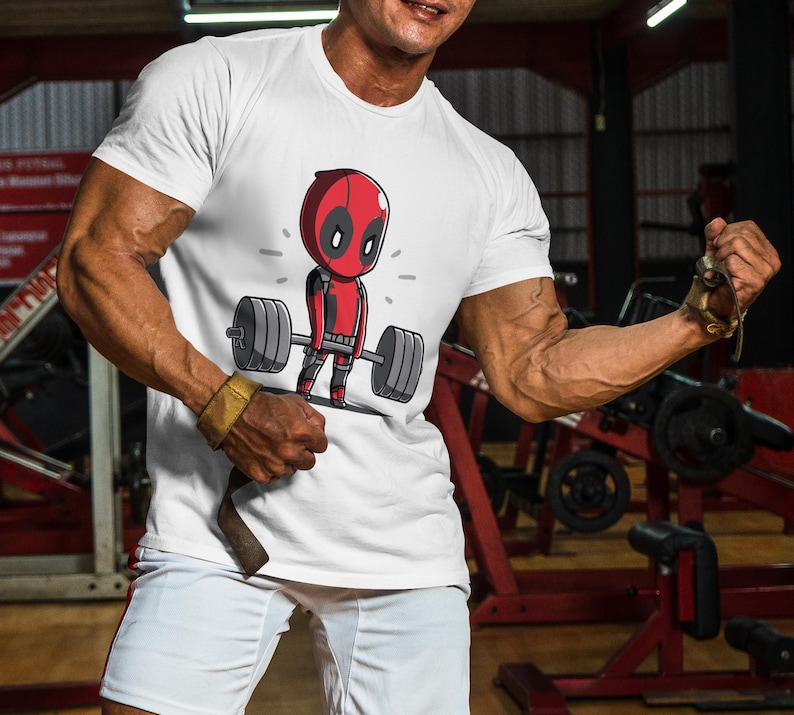 May include: White t-shirt featuring a cartoon image of Deadpool lifting weights. The image is in black, red, and white.
