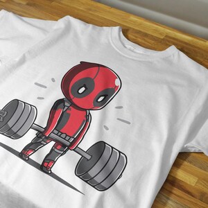 May include: White t-shirt featuring a cartoon image of Deadpool lifting a barbell. Deadpool is wearing his trademark red and black suit.