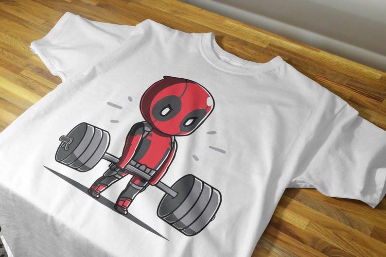 May include: White t-shirt featuring a cartoon image of Deadpool lifting a barbell. Deadpool is wearing his trademark red and black suit.