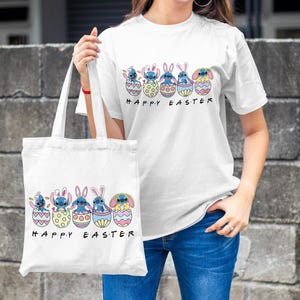 May include: White tote bag with a blue cartoon character holding an Easter egg. The bag has the text "Happy Easter" printed on it. A person is holding the bag and wearing a white t-shirt with the same design and the text "Happy Easter".